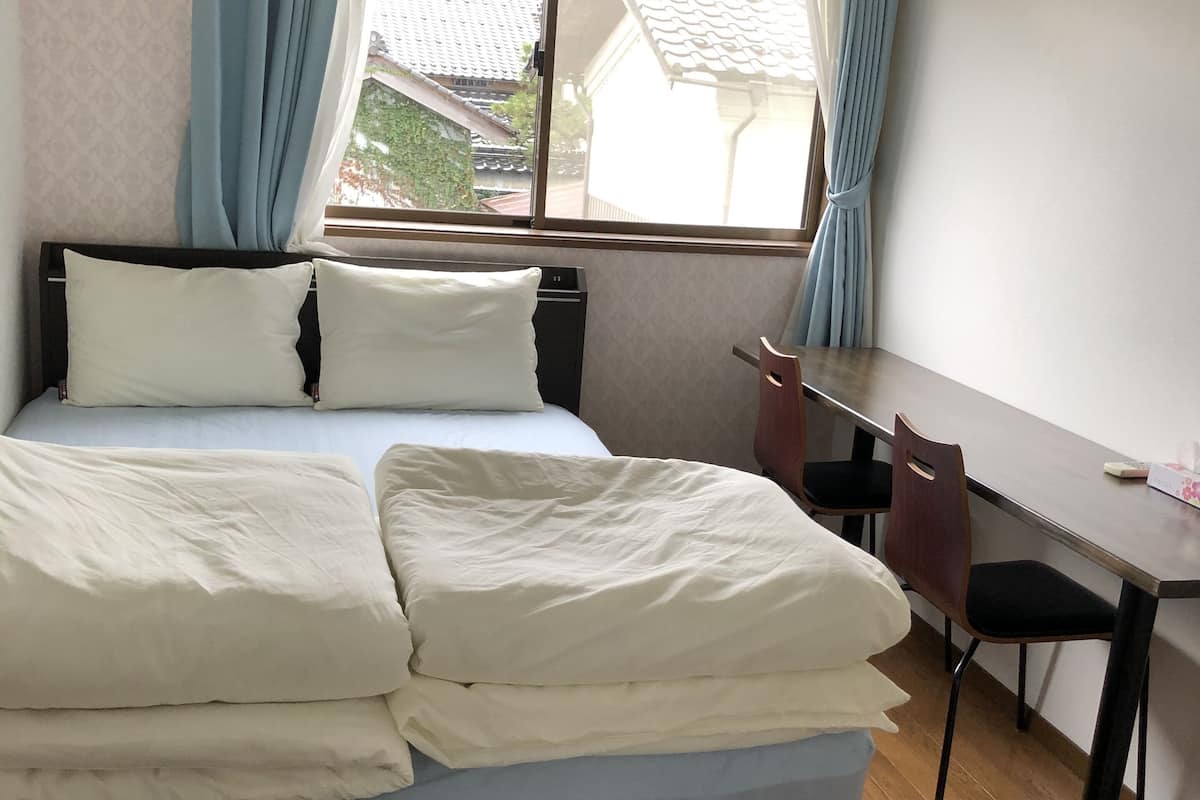 Una cama bien tendida con ropa blanca, un escritorio de madera con dos sillas y una ventana con cortinas azules.