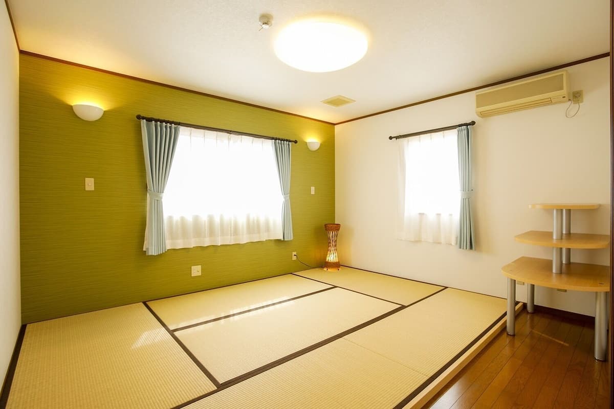 Una habitación con suelo de tatami, dos ventanas con cortinas, una pequeña mesa y aire acondicionado instalado en la pared.