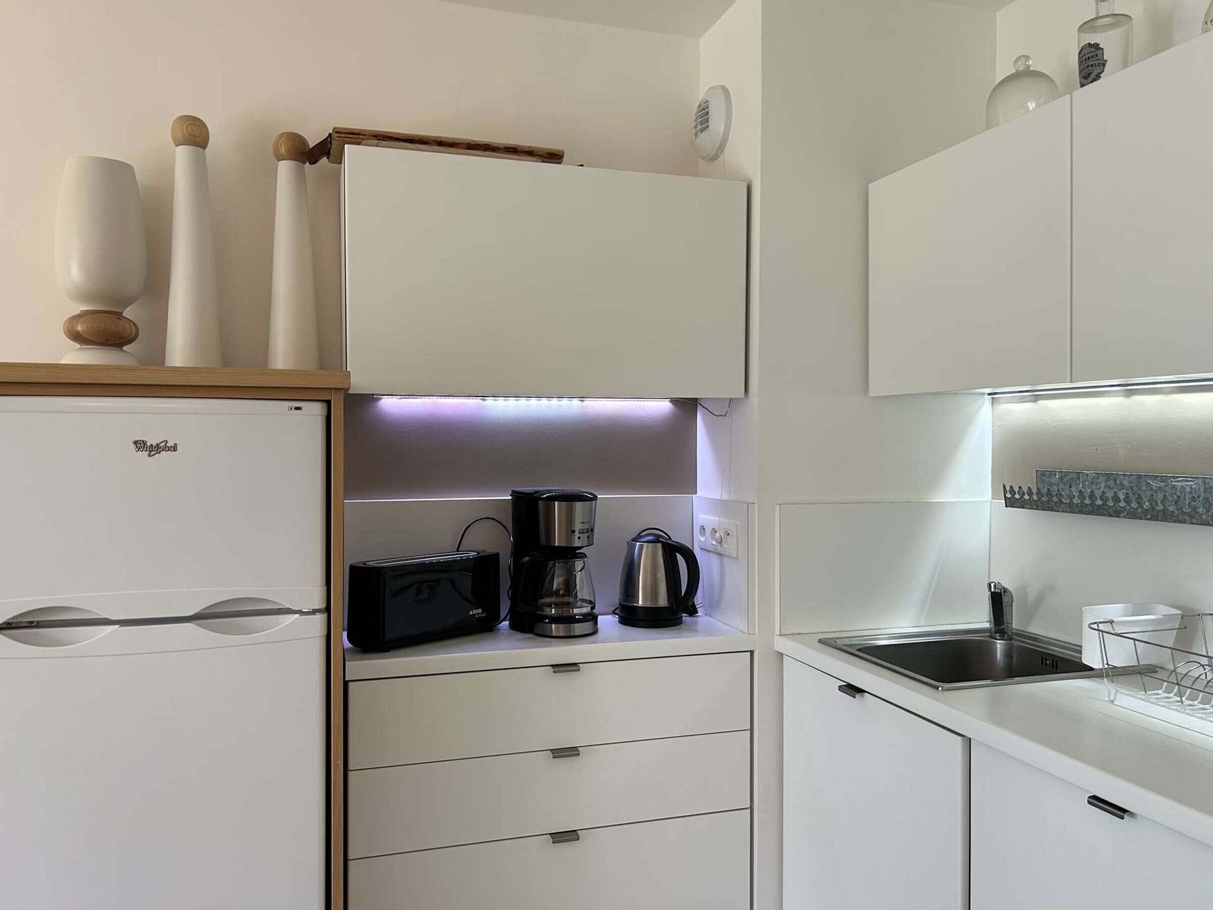 Una cocina compacta con muebles blancos, un refrigerador Whirlpool, una cafetera y un tostador.