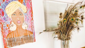 Ein Zimmer mit einem Korbssessel, einem Glastisch, einem Wandbild und einer Vase mit getrockneten Blumen.