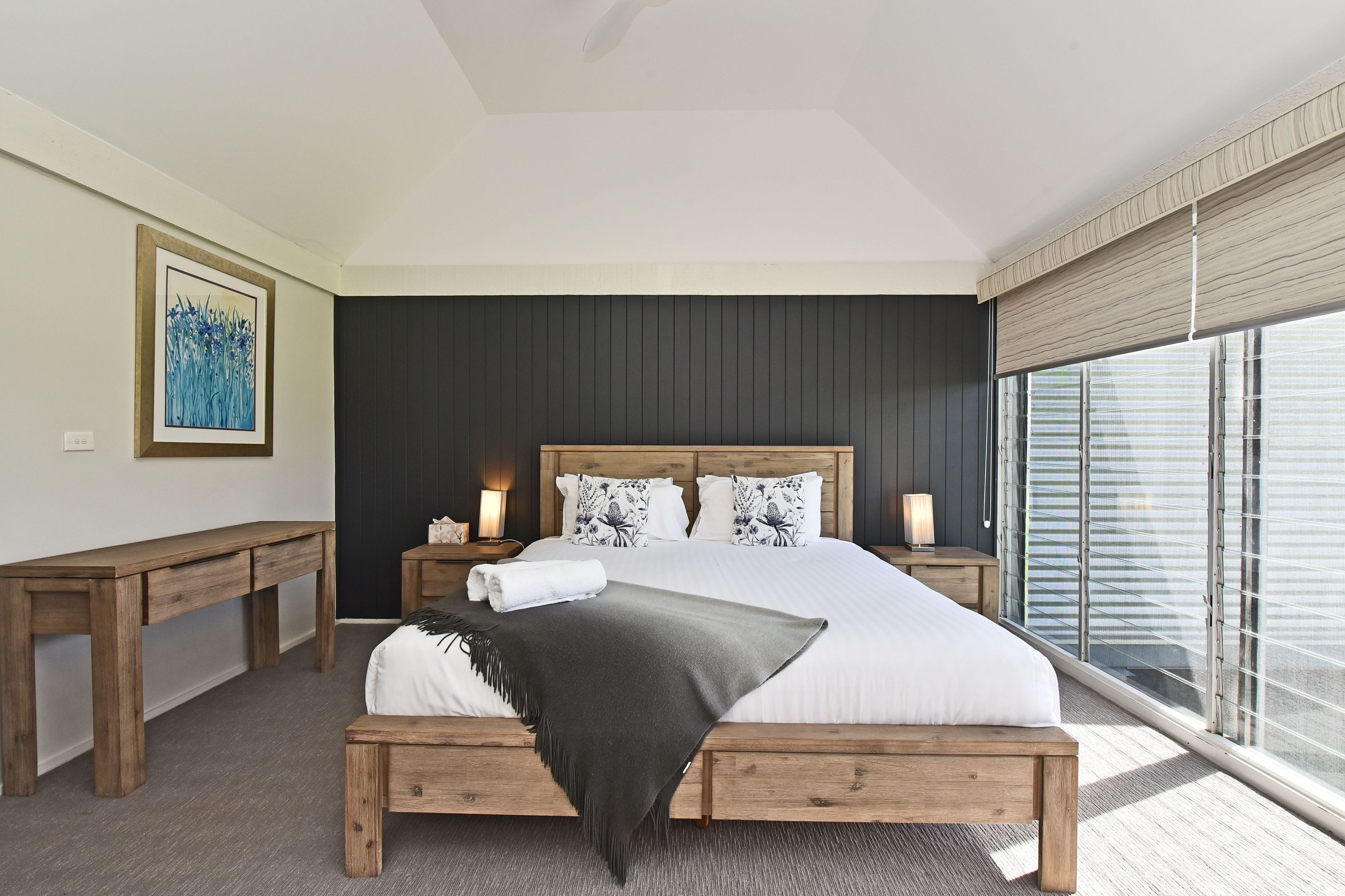 A modern bedroom with a large bed, wooden nightstands, a bedside table, and a window with blinds.