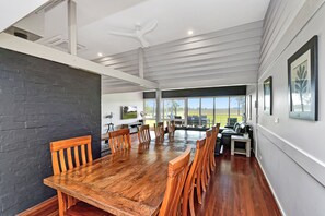 A spacious dining area with a large wooden table and chairs, a ceiling fan, and a view of the outdoors through large windows.