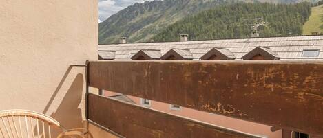 A balcony with a wicker chair, wooden railing, and a view of mountains and a village.