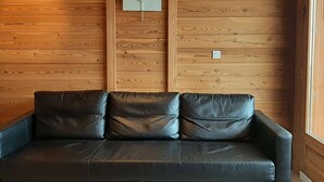 L’intérieur d’un chalet en bois, avec un canapé en cuir noir adossé à un mur en bois, un interrupteur et une fenêtre sur la droite.