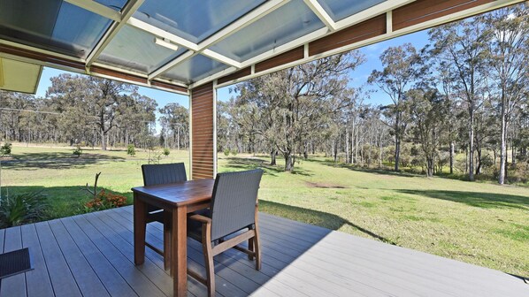 A covered outdoor area with a wooden table and chairs, overlooking a grassy landscape with trees.