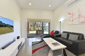 A modern living room with a flat-screen TV, a black leather sofa, a white coffee table, and a large window with a view of a grassy field and trees.