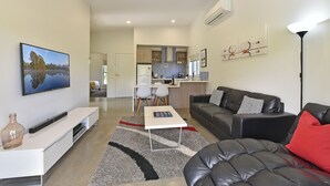 Ein modernes Wohnzimmer mit Flachbildfernseher, einer Couch, einem Couchtisch und einem Essbereich, im Hintergrund ist eine Küche erkennbar.