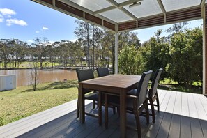 A covered outdoor seating area with a table and chairs, overlooking a body of water and a grassy landscape.