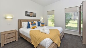 Ein Schlafzimmer mit Bett, Nachttischen, einer Wandleuchte, einem Fenster mit Jalousien und Blick ins Grüne.