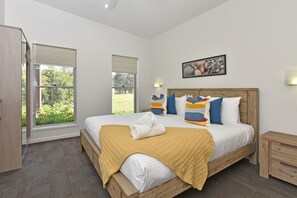 A bedroom with a large bed, a wooden headboard, a window with blinds, a bedside table, and a wall-mounted picture.