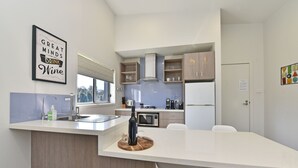 A modern kitchen with a central island, a dining area with chairs, and a kitchen counter with a wine bottle and glasses. - Ironbark Hill Spa Bath Villa 4 - Novato (Pokolbin)