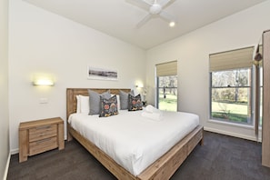 A bedroom with a large bed, a ceiling fan, a window with a view of the outdoors, and a small bedside table with a lamp.