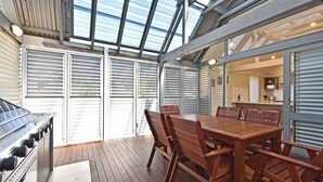 A covered outdoor dining area with a wooden table and chairs, a grill, and a view through the open shutters. - Ironbark Hill Estate (Pokolbin)