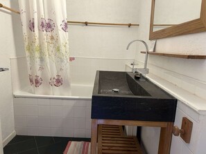 Un baño con un lavamanos negro, un tocador de madera, una bañera blanca con cortina de ducha estampada con flores y un estante de madera sobre el lavamanos.