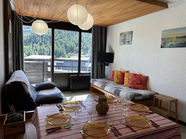 Una habitación acogedora con sofá, una mesa de comedor dispuesta con platos y vasos, y vista a un bosque a través de grandes ventanales.