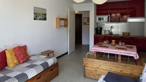A cozy room with a wooden ceiling, a bed with a checkered blanket, a wooden bench, a dining table with a striped tablecloth, and a kitchen area with red cabinets and a microwave. - Cozy 2-Room Apartment with Mountain View, Balcony, and Parking in Montgenèvre - Sleeps 6 (Montgenèvre)