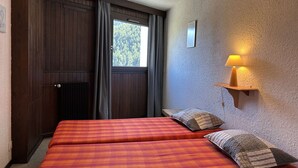 A room with two beds, a window with curtains, a wall-mounted lamp, and a wooden door. - Cozy 2-Room Apartment with Mountain View, Balcony, and Parking in Montgenèvre - Sleeps 6 (Montgenèvre)