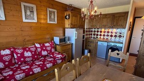 A cozy kitchen with wooden cabinets, a microwave, a refrigerator, and a dining table with a red patterned sofa. - 2-room rental for 5 people at the foot of the slopes (Les Adrets-Prapoutel)