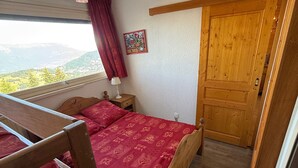 A bedroom with a wooden bed, a nightstand, a lamp, a window with a view, and a door. - 2-room rental for 5 people at the foot of the slopes (Les Adrets-Prapoutel)