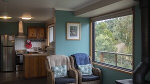 A kitchen with a central island, wooden cabinets, a stainless steel refrigerator, and a stove. There is a wicker chair with cushions and a large window with a view of greenery.