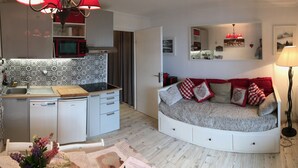 Un espace de vie compact comprenant un coin cuisine, un canapé-lit avec des coussins rouges, une table à manger décorée de fleurs et un lampadaire.