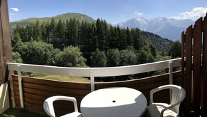 Un balcon avec une balustrade blanche, deux chaises en plastique blanches et une petite table ronde. On aperçoit, au loin, une chaîne de montagnes boisée.