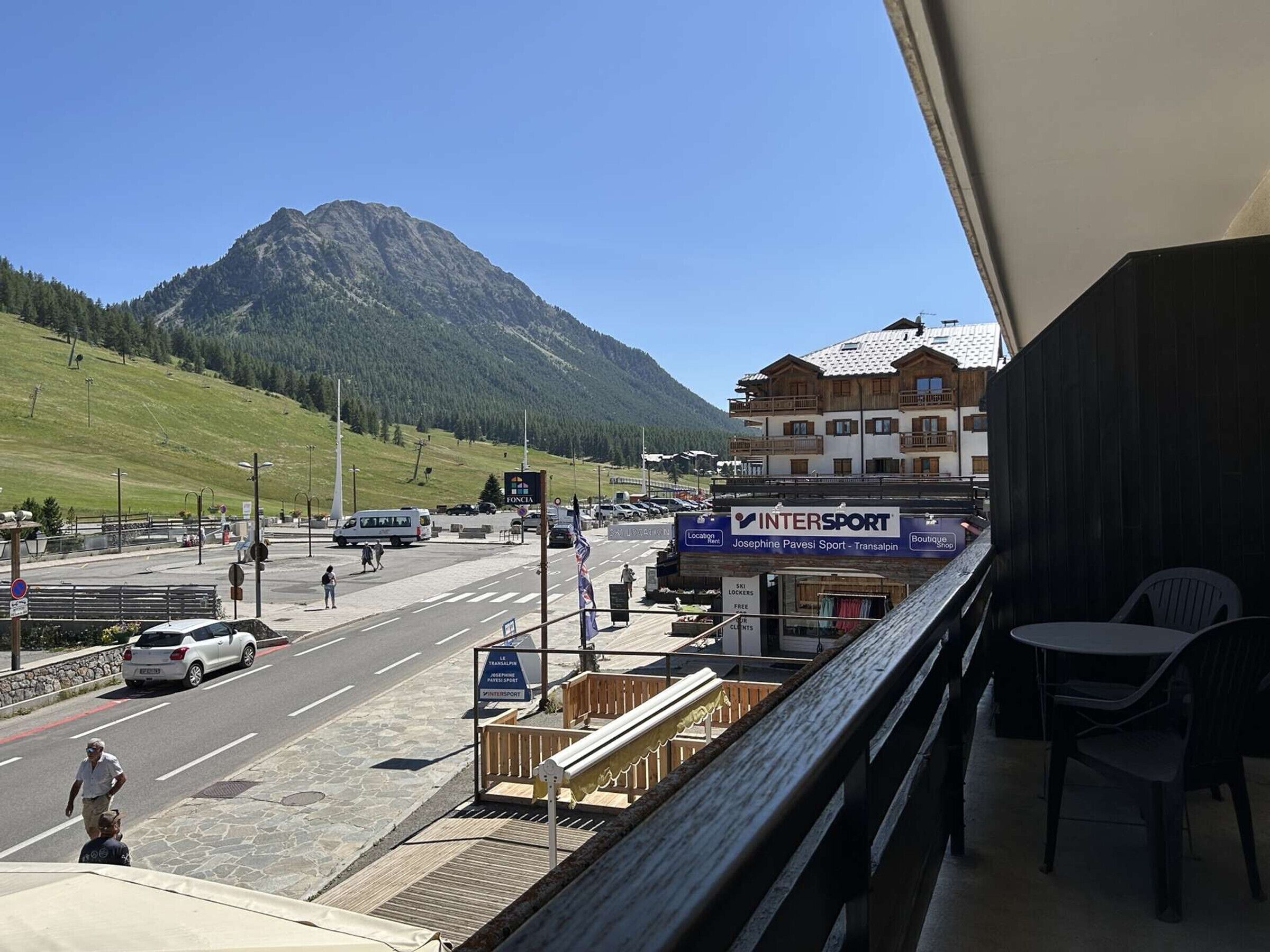 A view from a balcony overlooking a street with a mountain in the background, a sign for 'INTERSPORT' with a boutique shop, and a 'FONCIA' sign.