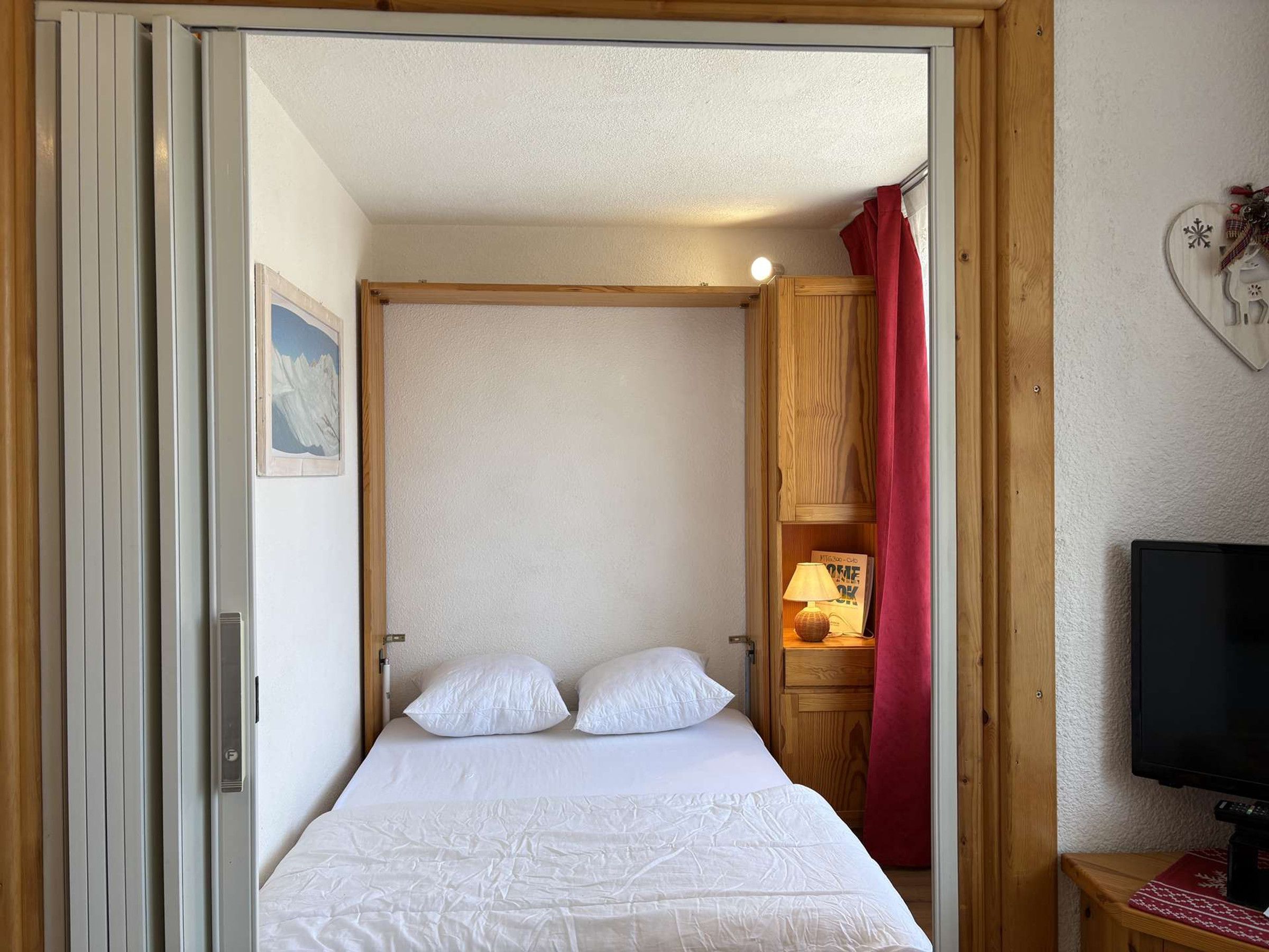 A bedroom with a bed, a nightstand with a lamp, a television, and a red curtain.