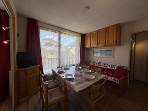A dining area with a table set for four, wooden cabinets, a television, and a view of buildings through the window. - Apartment Montgenèvre - 2 bedrooms, balcony, washing machine, TV (Montgenèvre)