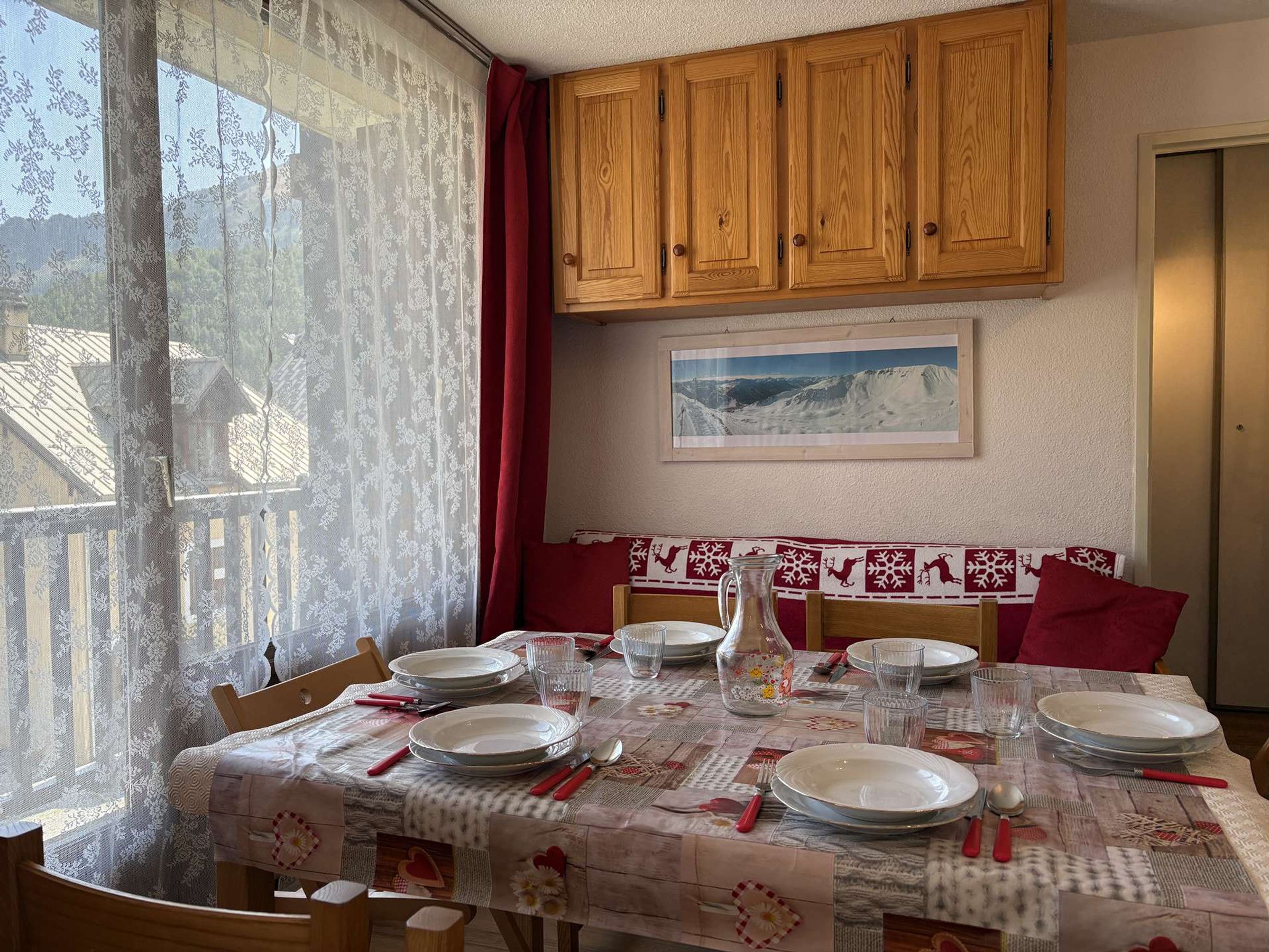 Un comedor con una mesa dispuesta para cuatro, armarios de madera arriba, un ventanal con cortinas de encaje y un cuadro en la pared.