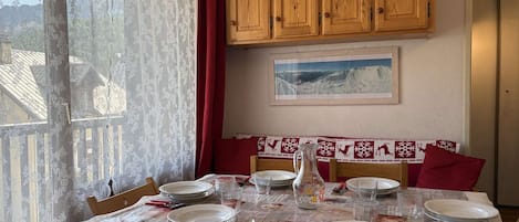 Un comedor con una mesa dispuesta para cuatro, armarios de madera arriba, un ventanal con cortinas de encaje y un cuadro en la pared.