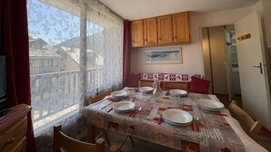 A dining area with a table set for four, wooden cabinets above, a window with lace curtains, and a door leading to another room.