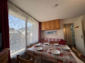 Un coin repas avec une table dressée pour quatre personnes, des placards en bois au-dessus, une fenêtre ornée de rideaux en dentelle et une porte donnant sur une autre pièce.