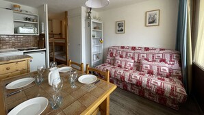 Un espace de vie compact comprenant une table à manger pour quatre personnes, un coin cuisine, un canapé et une petite bibliothèque.