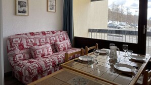 Une pièce chaleureuse avec un canapé à motifs, une table à manger dressée avec des assiettes, des verres et des couverts, et une vue sur un parking par une grande fenêtre.