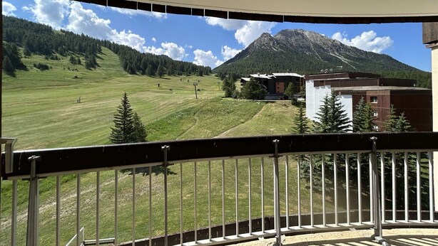 Un balcón con vista a una montaña, una pista de esquí y un edificio con techo rojo.