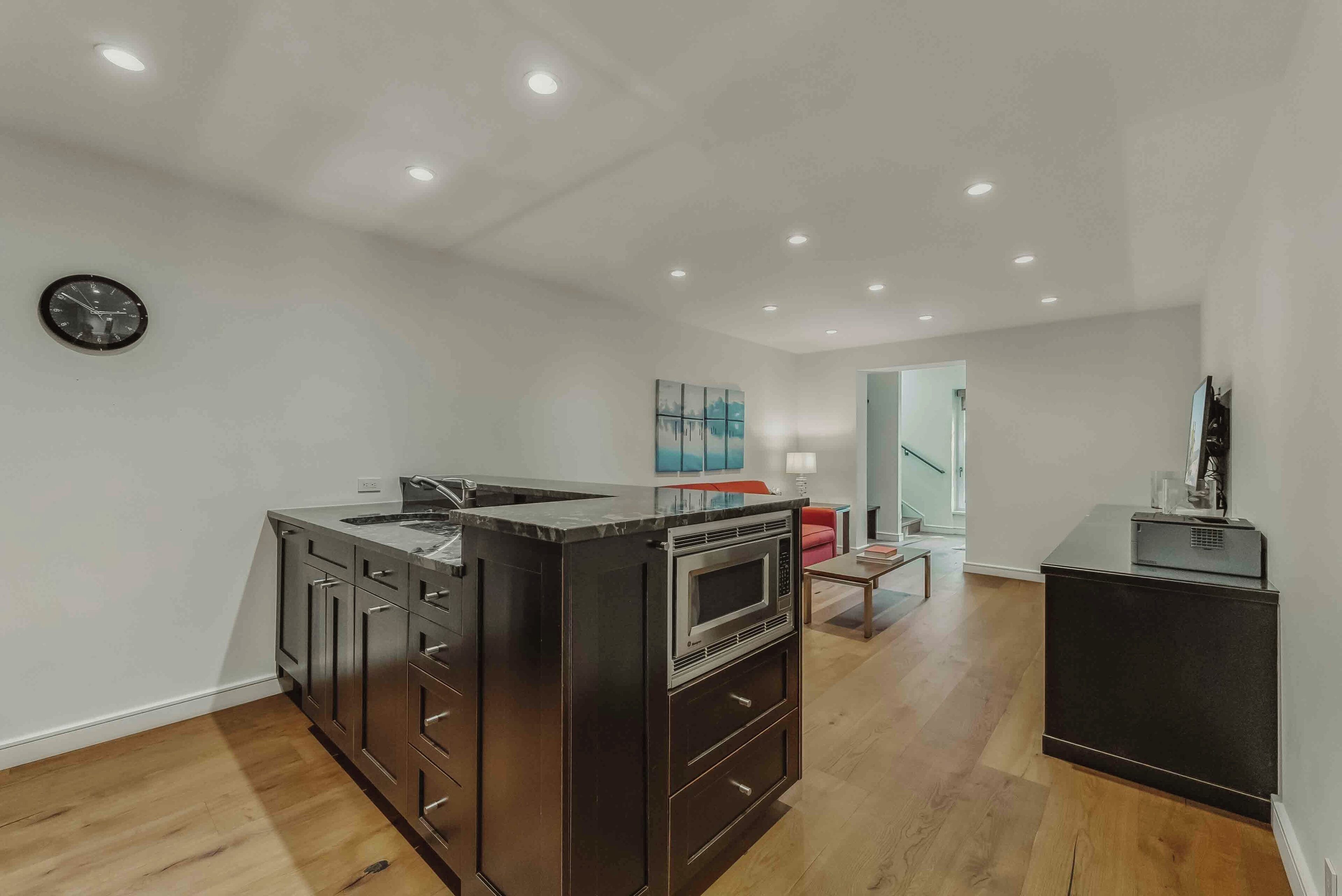 A modern kitchen with dark cabinetry, a built-in microwave, and a sink. There is a clock on the wall and a painting above the counter.