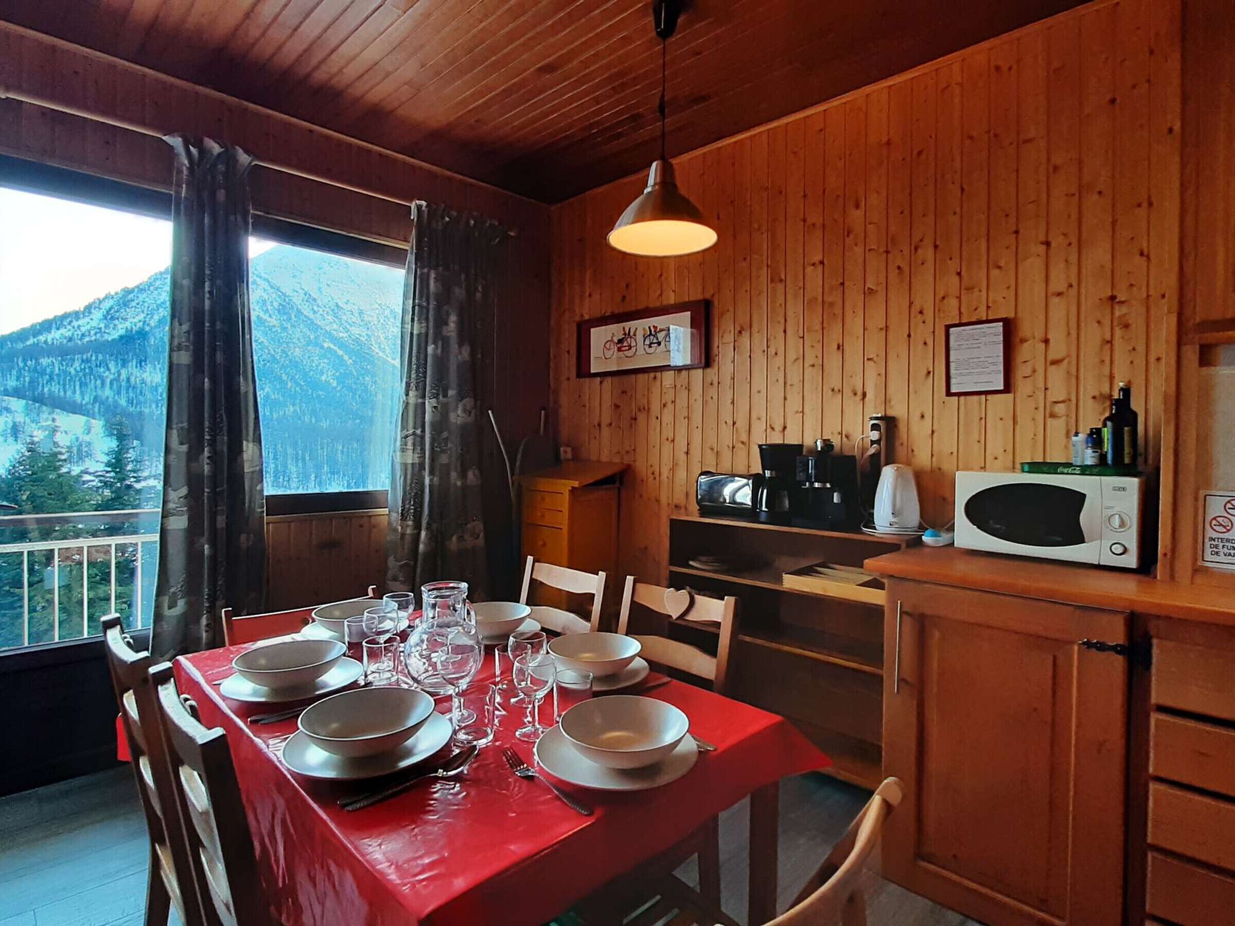 A wooden cabin with a dining table set for four, kitchen appliances, and a view of mountains.
