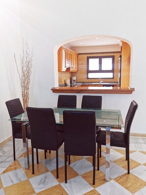 A dining area with a glass table and black chairs, a kitchen with wooden cabinets and a window above the sink, and a decorative plant.