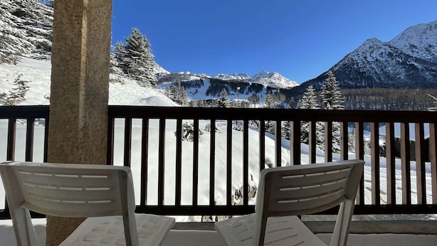 Een balkon met twee witte stoelen, een houten leuning en uitzicht op een besneeuwde berg.