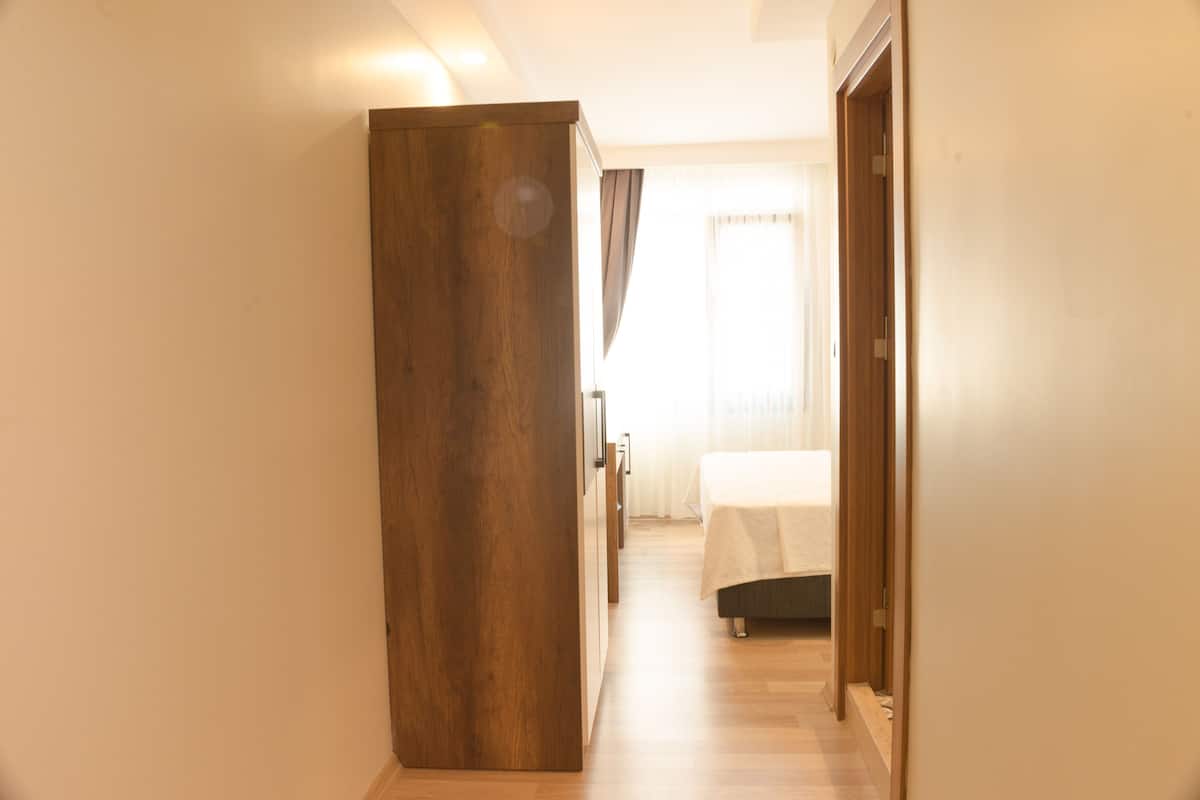 A hallway leading to a bedroom with a bed, a wooden wardrobe, and a window with curtains.