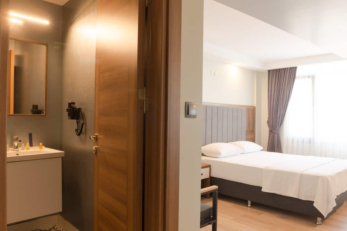 A hotel room with a wooden door, a bedside table, a bed with white bedding, a nightstand, a mirror, and a window with curtains.