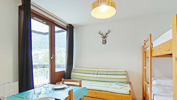 A room with a bunk bed, a table set for two, a window with curtains, and a wall-mounted deer head decoration.