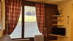 A living room with a sofa, a TV on a shelf, a radiator, and a window with curtains. - Ski-in/Ski-out 6 Pax Apartment, Montgenèvre with East Balconies and Modern Amenities (Montgenèvre)