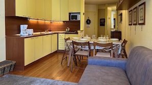 A compact kitchen with yellow cabinets, a microwave, and a coffee maker. There is a dining area with a table set for four and a blue sofa. - Ski-in/Ski-out 6 Pax Apartment, Montgenèvre with East Balconies and Modern Amenities (Montgenèvre)