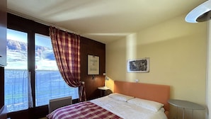 A hotel room with a bed, a small table, a lamp, a window with curtains, and a painting on the wall. - Ski-in/Ski-out 6 Pax Apartment, Montgenèvre with East Balconies and Modern Amenities (Montgenèvre)
