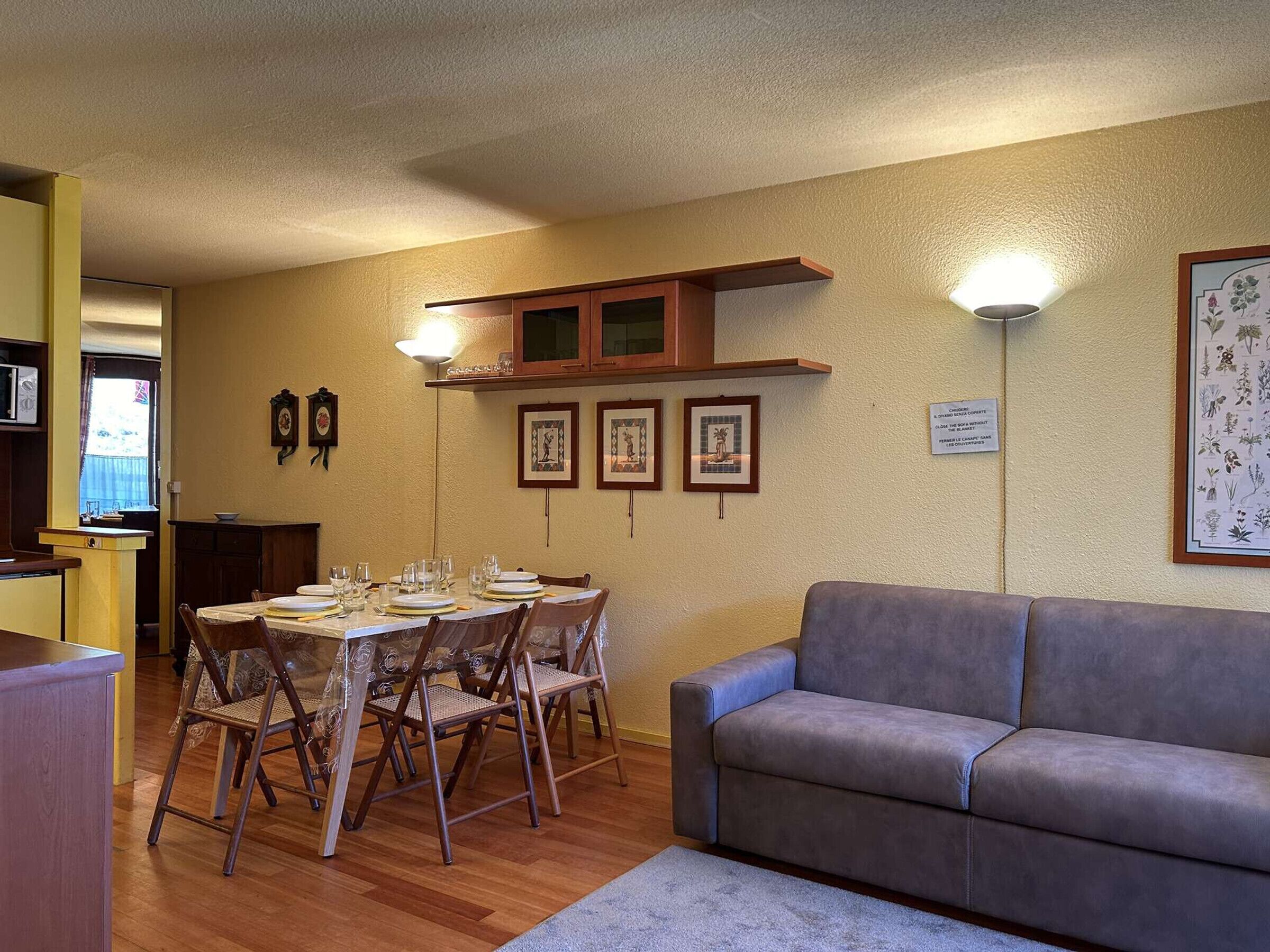A dining area with a table set for four, a grey sofa, and framed botanical prints on the wall.