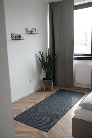 A modern bedroom with a wooden floor, a grey rug, a bed, a potted plant, and two wall-mounted shelves with small plants.