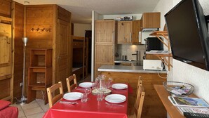 A compact kitchen with wooden cabinets, a dining table set for four, and a television mounted on the wall. - Modern studio PMR, south balcony sleeps 4 - Montgenèvre (Montgenèvre)