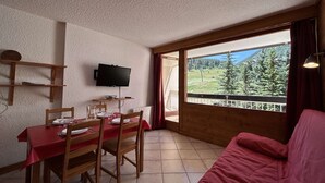 A dining area with a table set for four, a television mounted on the wall, and a large window offering a view of a forested landscape. - Modern studio PMR, south balcony sleeps 4 - Montgenèvre (Montgenèvre)